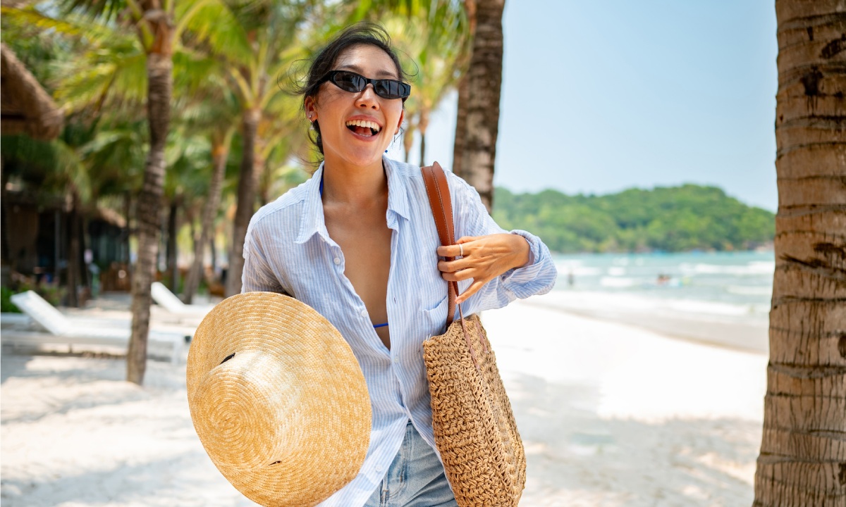 Mujer con gafas de sol, sombrero y bolso de playa en playa de palmeras