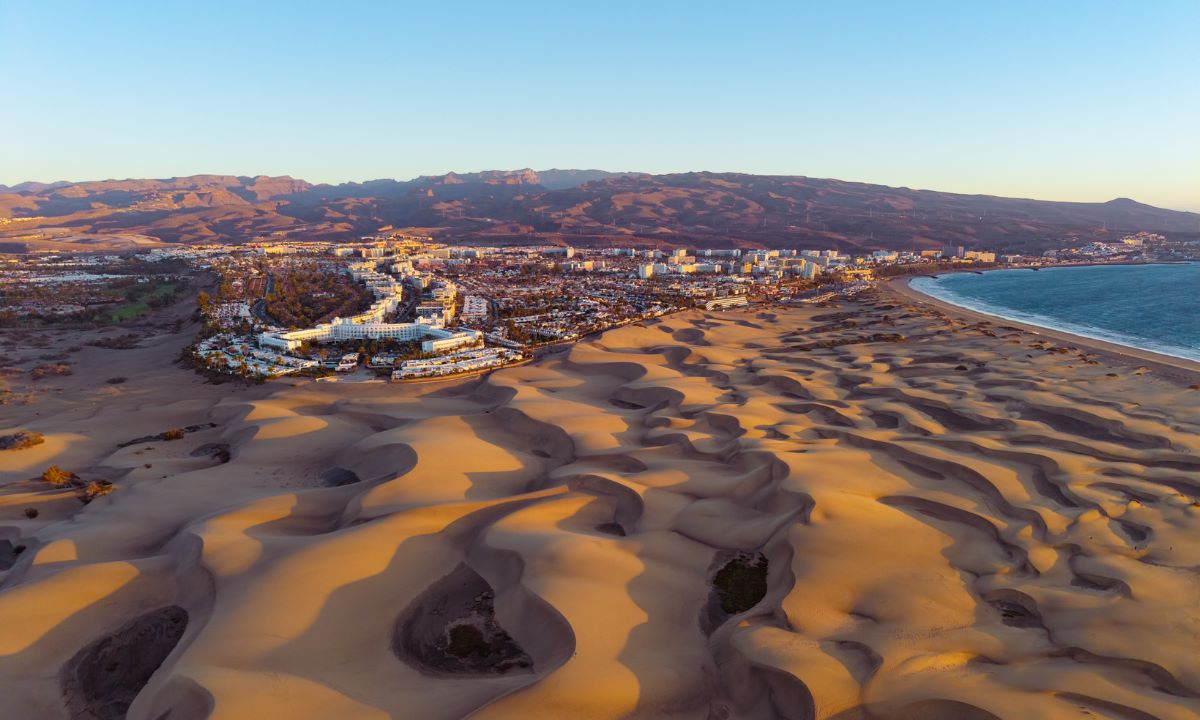 Aerial view of the spectacular Maspalomas Dunes in Gran Canaria at sunset, with hotels and mountains in the background.