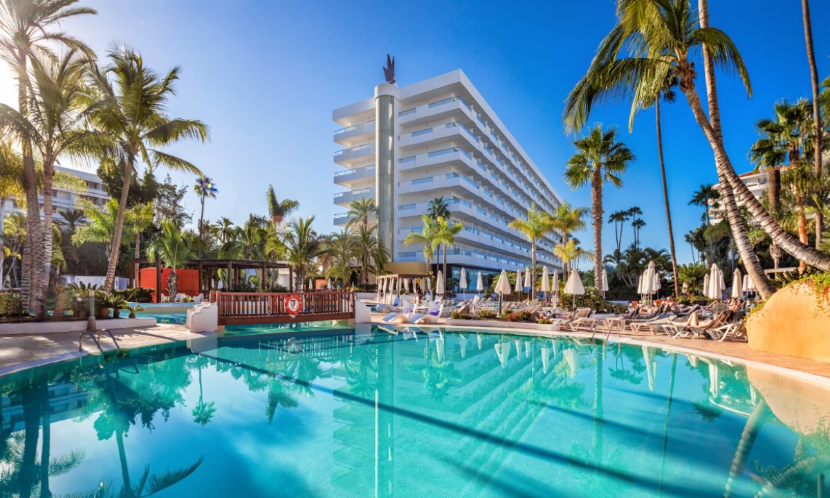 Main swimming pool with crystal-clear water at the Gran Canaria Princess hotel, surrounded by palm trees and sun loungers under a clear sky.